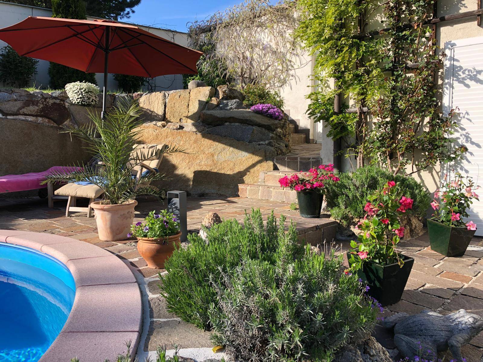 Entspannungsbereich am Pool mit Felsenlandschaft, mediterranen Pflanzen und Sonnenschirm.