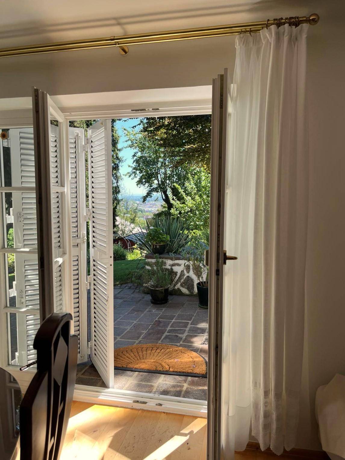 Blick aus dem Innenraum durch offene Fenstertüren auf die gepflasterte Terrasse und den Garten.