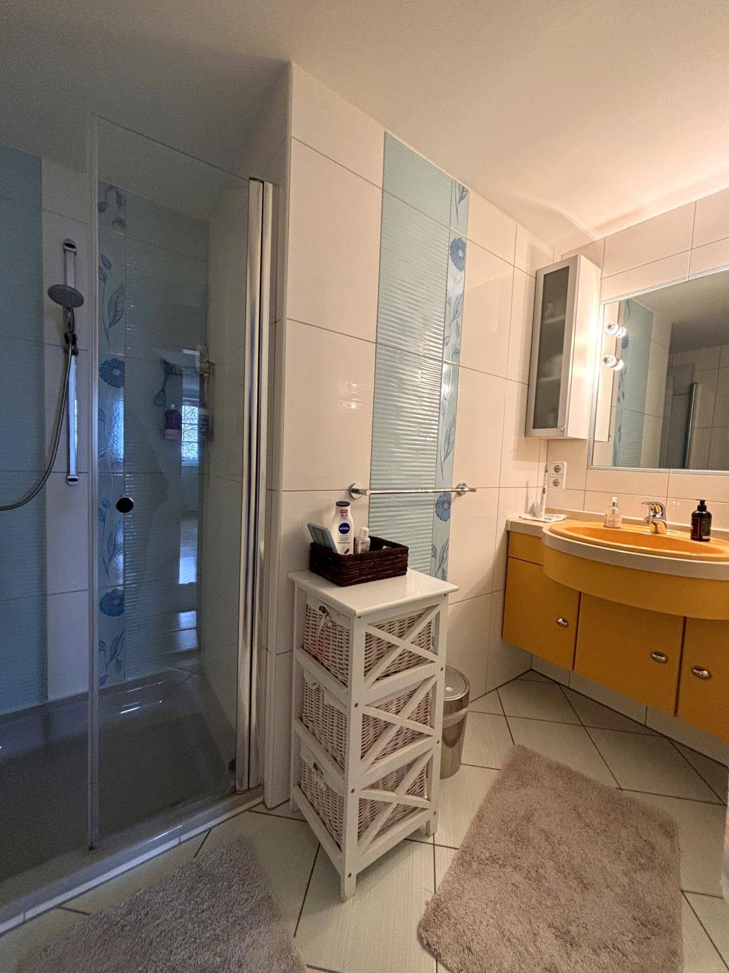 Badezimmer mit Duschkabine, gelbem Waschbeckenunterschrank und hellen Fliesen an Wänden und Boden.