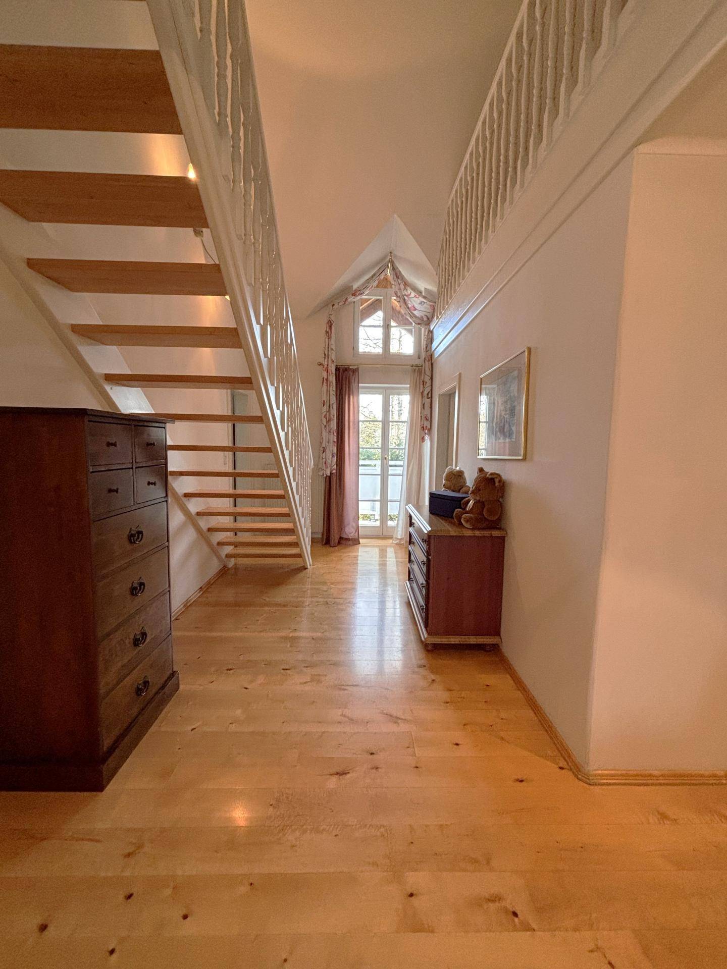 Heller Flur mit Holztreppe, Holzboden und hohen Decken, die viel Licht hereinlassen.