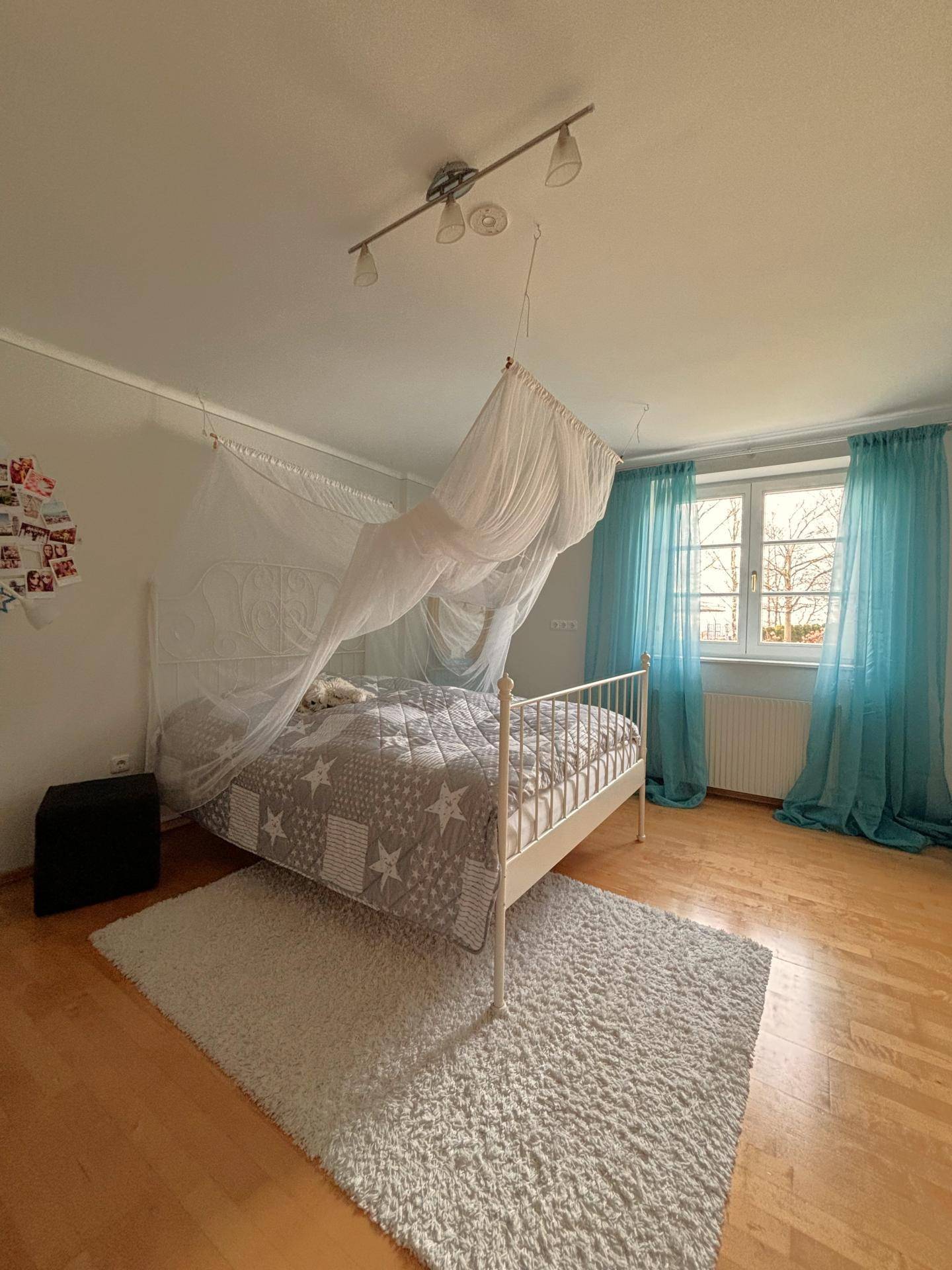Helles Schlafzimmer mit Holzboden, weißem Metallbett und auffälligen türkisen Vorhängen am Fenster.