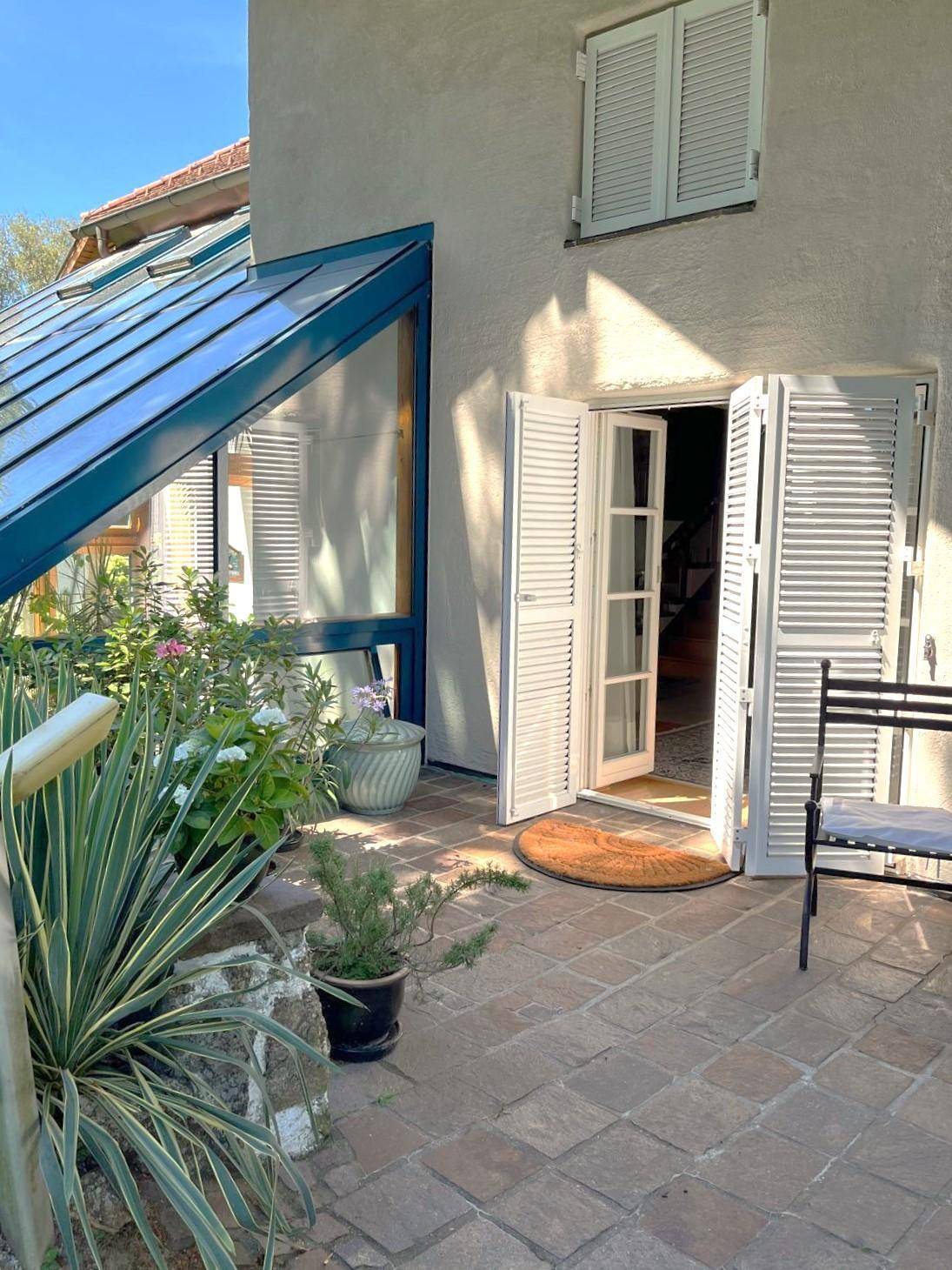 Eingangsbereich des Hauses mit gepflasterter Terrasse, offenen Türen und Wintergarten-Anbau.