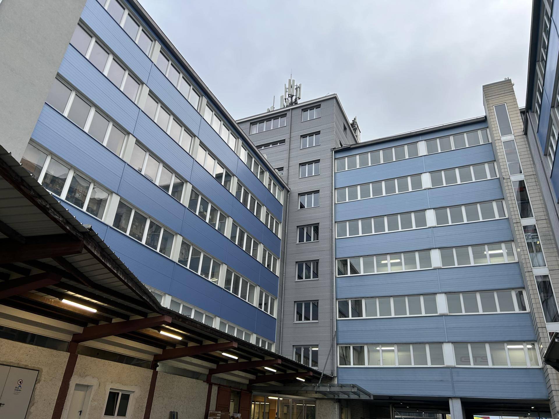 Mehrere Gebäudeteile mit blauen und grauen Fassaden unter bewölktem Himmel.