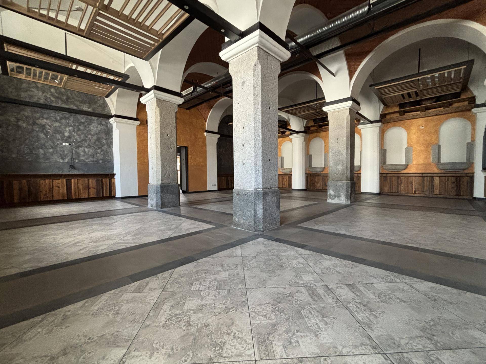 Weitläufiger, leerer Saal mit hohen Gewölbedecken, massiven Steinsäulen und einem gemusterten Fliesenboden.