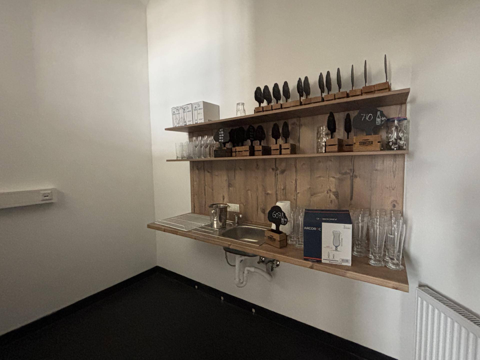 Kleine Küchenzeile mit Spülbecken, Holzregalen und Gläsern, ideal für einen Barbereich oder eine Teeküche.