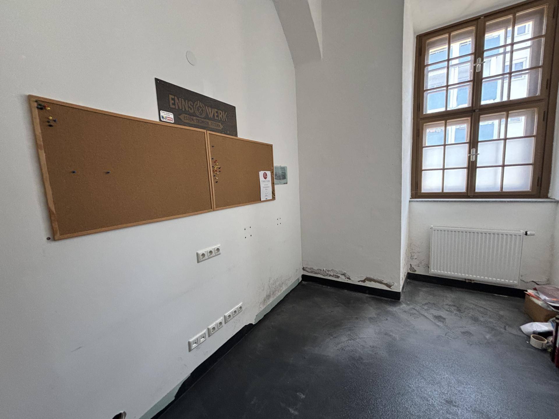 Kleiner Raum mit Fenster, Heizkörper und einer Pinnwand, geeignet als Büro oder Abstellraum.