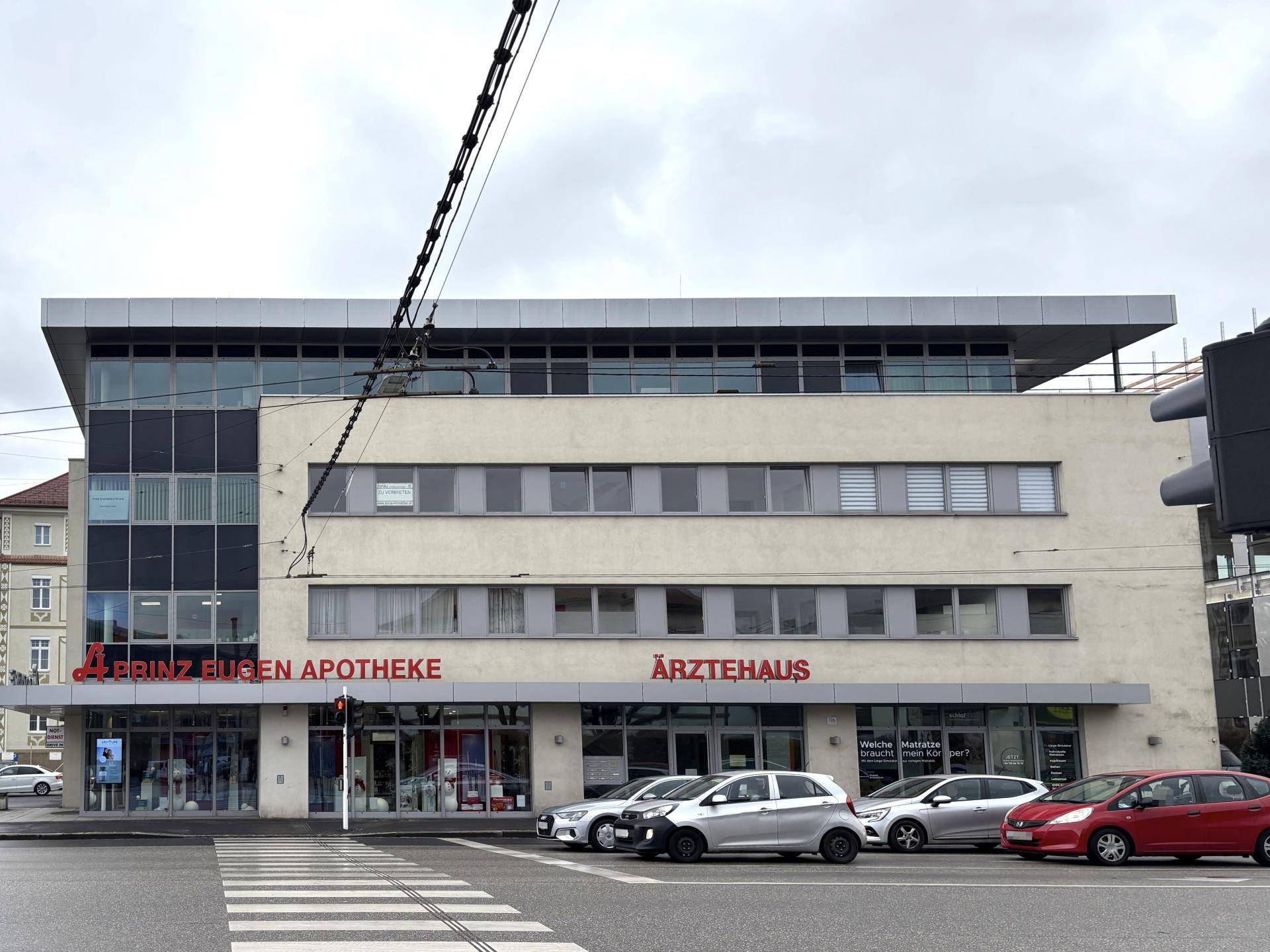 Ansicht des modernen Ärztehauses mit Geschäften im Erdgeschoss und parkenden Autos davor.