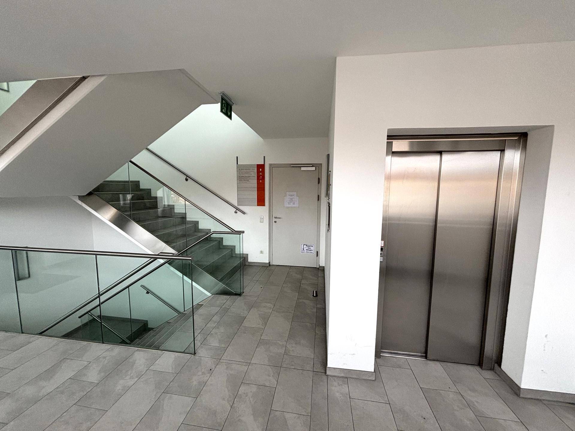 Moderner Eingangsbereich mit Treppe, Glasgeländer und einem Aufzug, der alle Etagen erschließt.