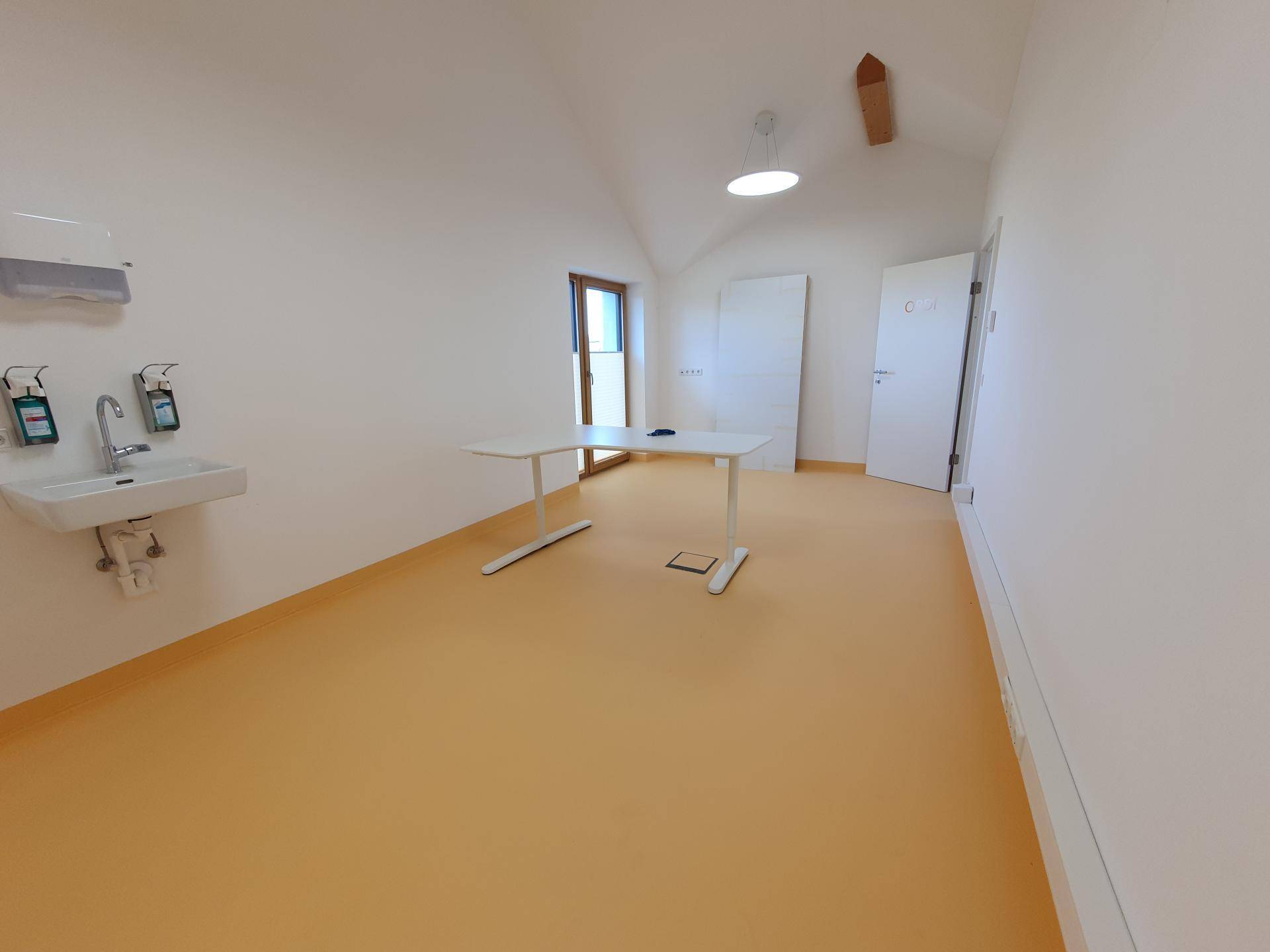 Heller Raum mit orangefarbenem Boden, weißen Wänden und einem Waschbeckenbereich. Ein Tisch und ein Fenster sind vorhanden.