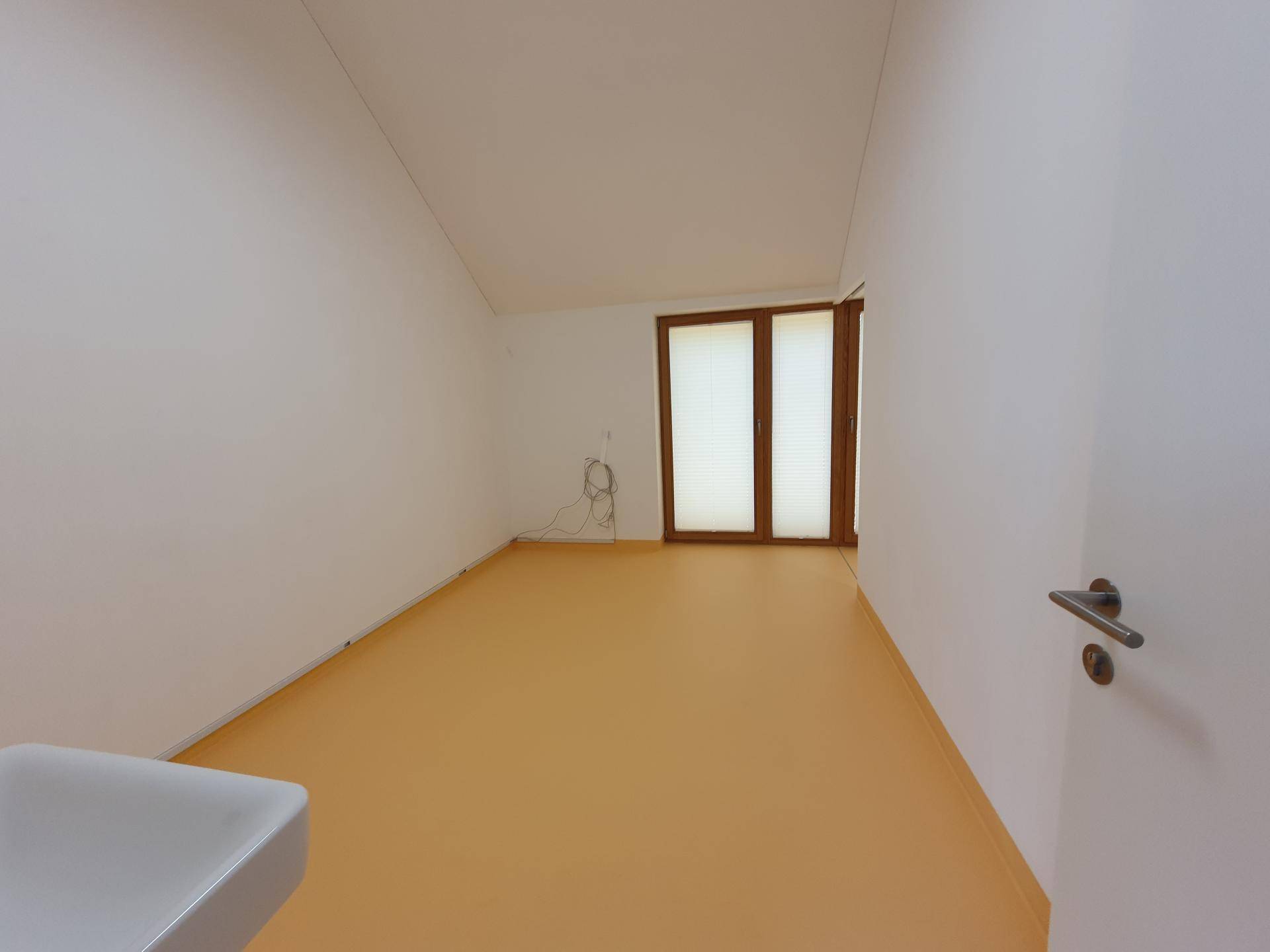 Heller Raum mit orangefarbenem Boden, weißen Wänden und einem Fenster mit Holzjalousien. Eine Tür ist rechts sichtbar.