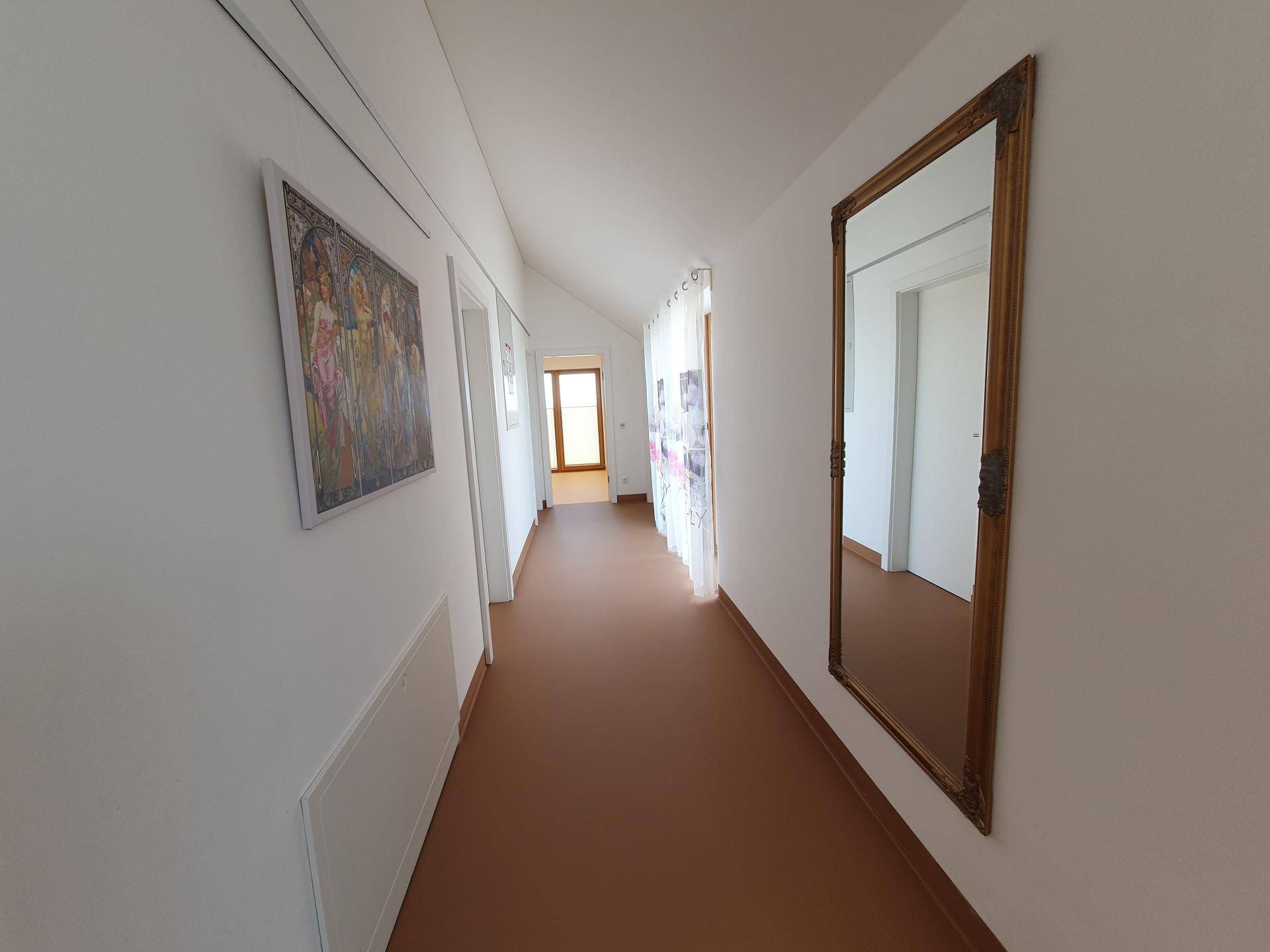 Langer, heller Flur mit weiß gestrichenen Wänden, braunem Boden und einem großen, verzierten Spiegel.