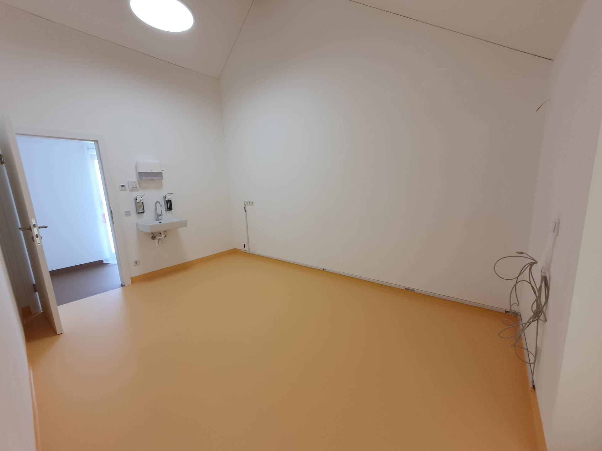Leerer Raum mit hellbraunem Boden, weißen Wänden und einem Waschbeckenbereich. Eine Tür und Kabel sind sichtbar.