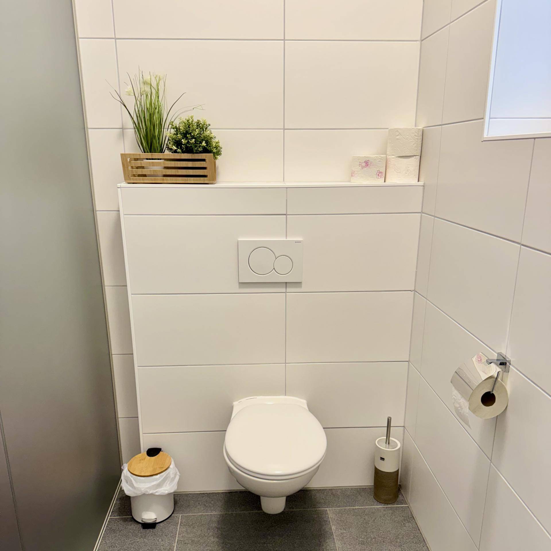 Saubere und moderne Toilette mit weißen Fliesen, Wand-WC und kleinem Fenster für Tageslicht.