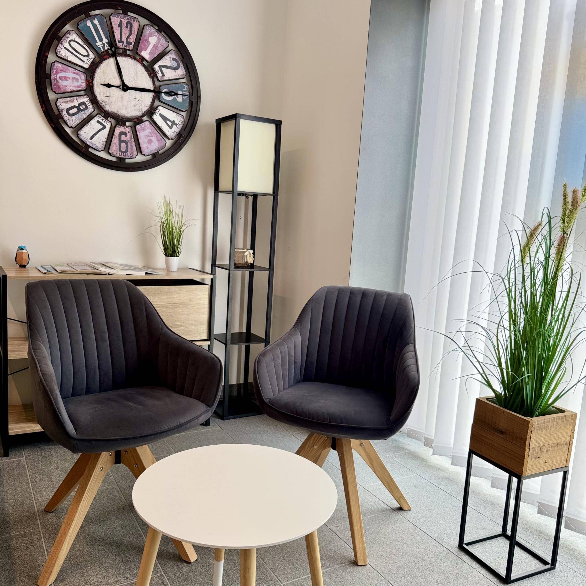 Gemütlicher Wartebereich mit zwei grauen Sesseln, einem kleinen Tisch und einem großen Wanduhr-Dekor.