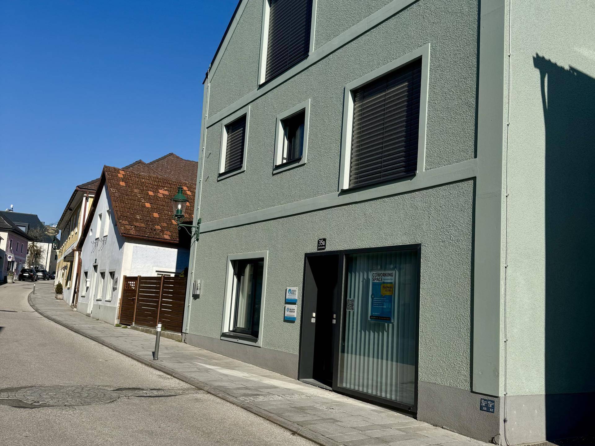 Modernes Gebäude mit hellgrüner Fassade, großen Fenstern und Rollläden an einer ruhigen Straße.