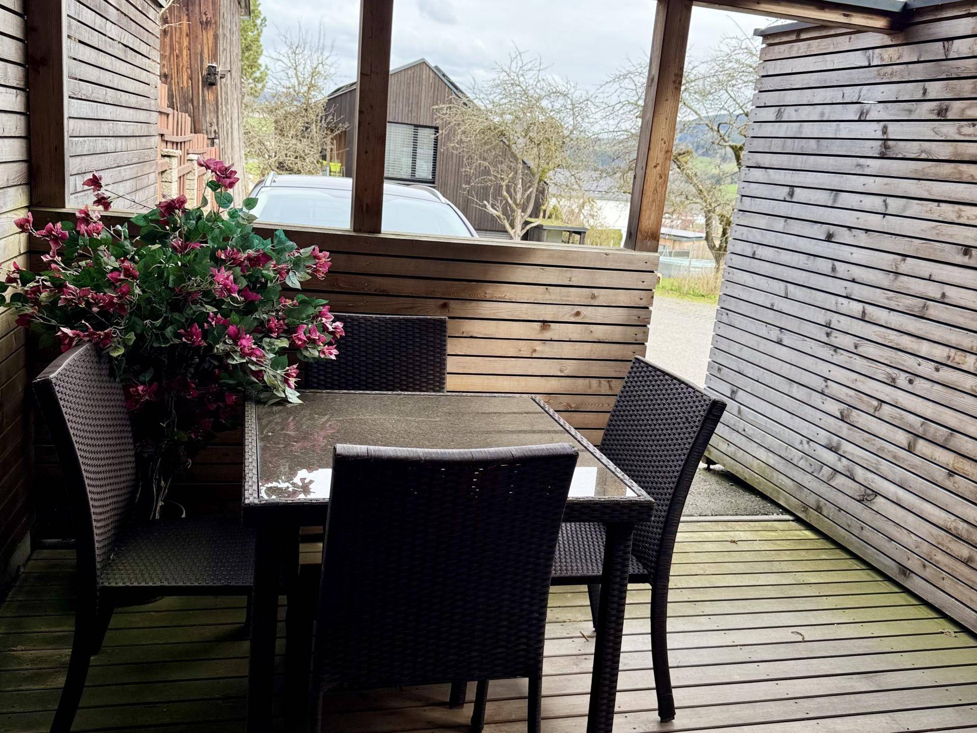 Überdachte Holzterrasse mit dunklen Korbmöbeln und Blick auf die Umgebung.