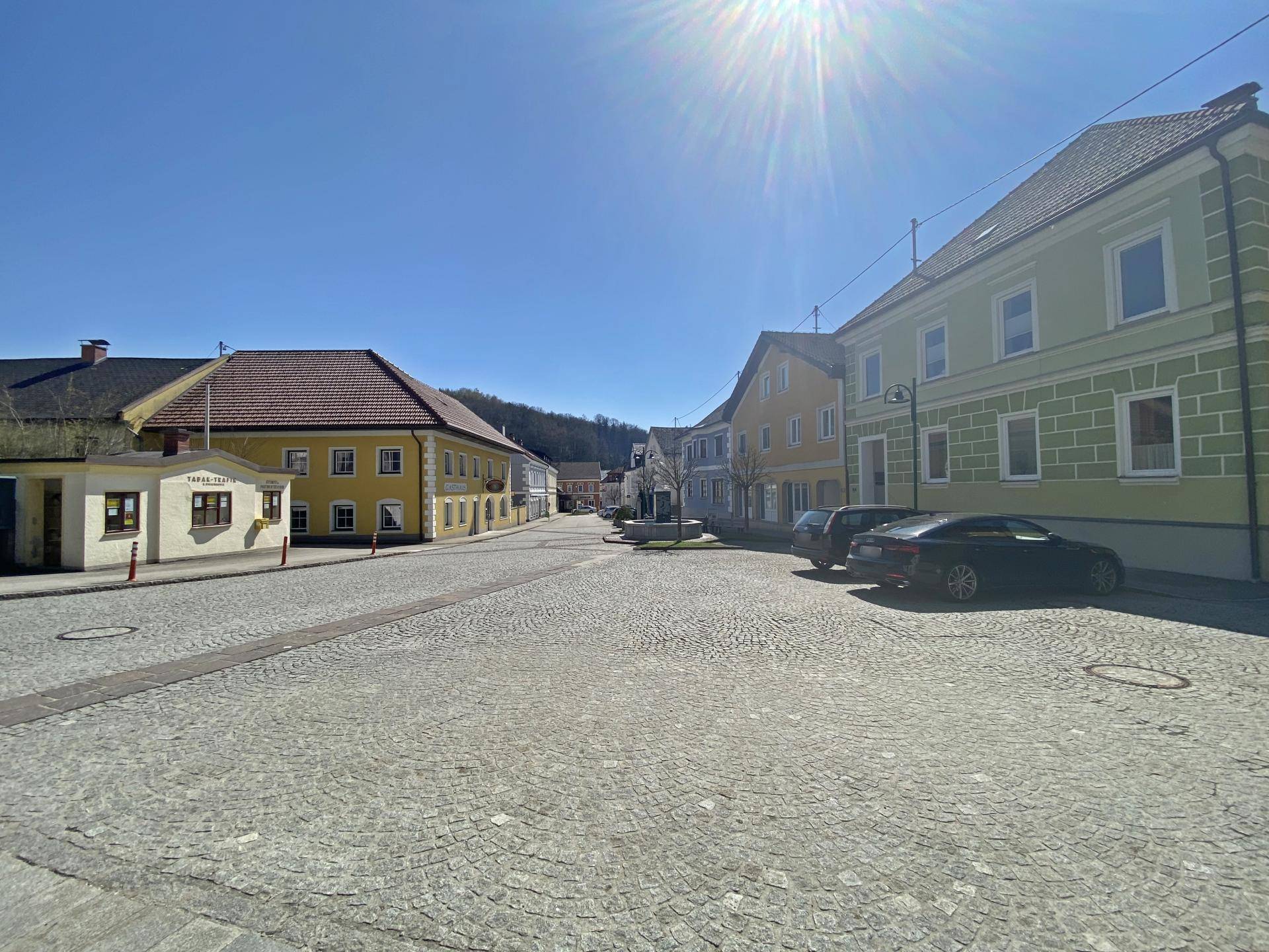 Sonniger Blick auf eine gepflasterte Dorfstraße mit traditionellen Gebäuden und parkenden Autos.