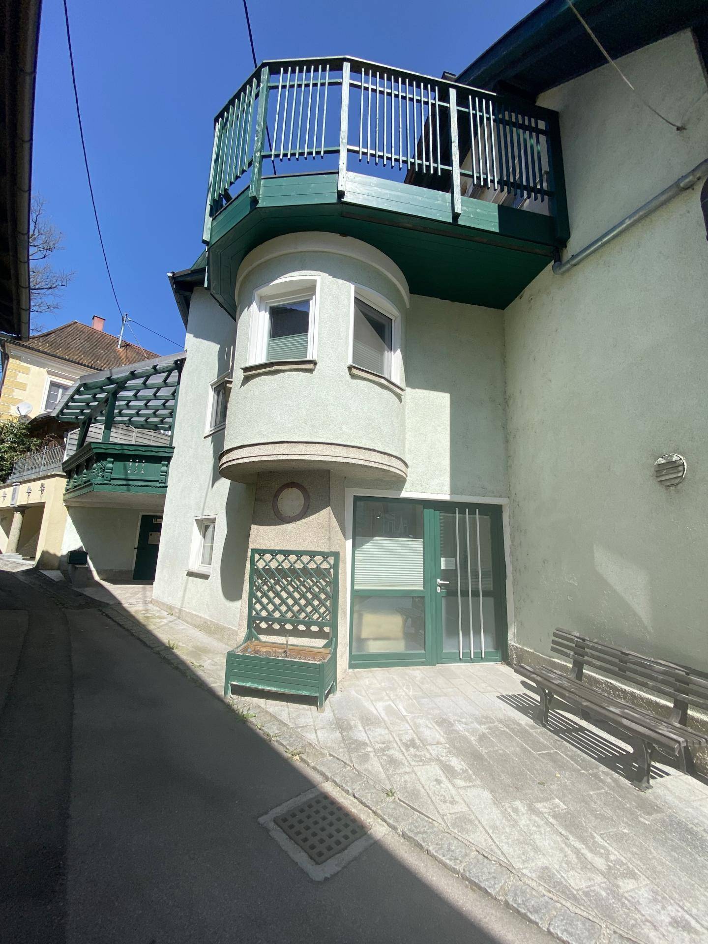 Seitenansicht eines Gebäudes mit grünem Balkon, Erker und Eingangsbereich an einem sonnigen Tag.