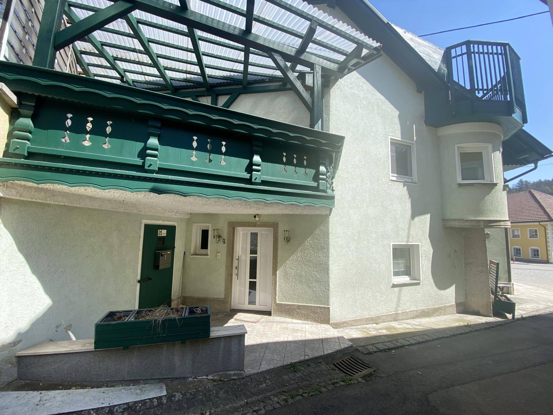 Vorderansicht des Hauses mit überdachter Terrasse, Haupteingang und grünem Balkon.
