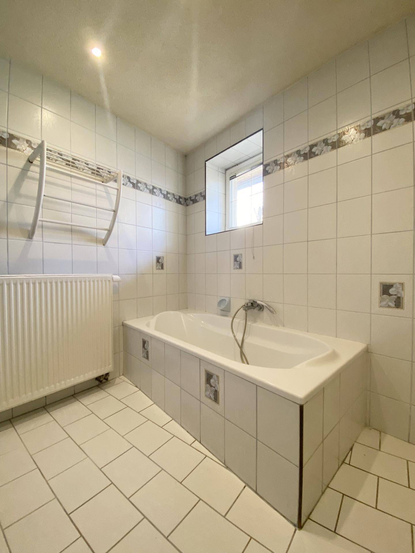 Badezimmer mit Badewanne, Heizkörper und weißen Fliesen, die ein klassisches Muster aufweisen.