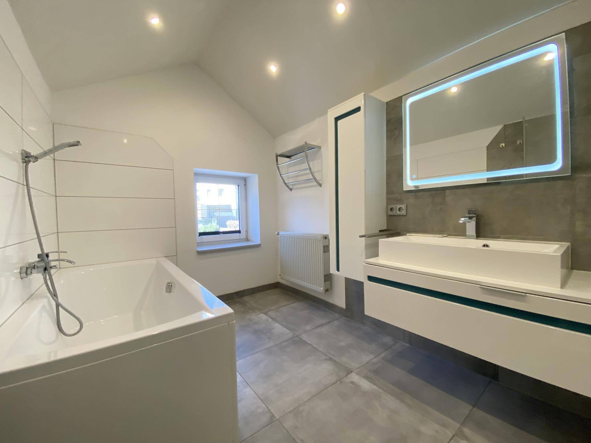 Modernes Badezimmer mit Badewanne, Waschtisch und beleuchtetem Spiegel, ausgestattet mit grauen Fliesen.
