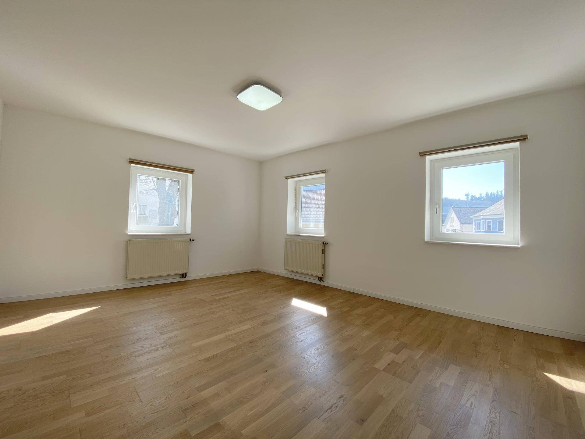 Helles Schlafzimmer mit Holzboden und drei Fenstern, die viel Tageslicht hereinlassen.
