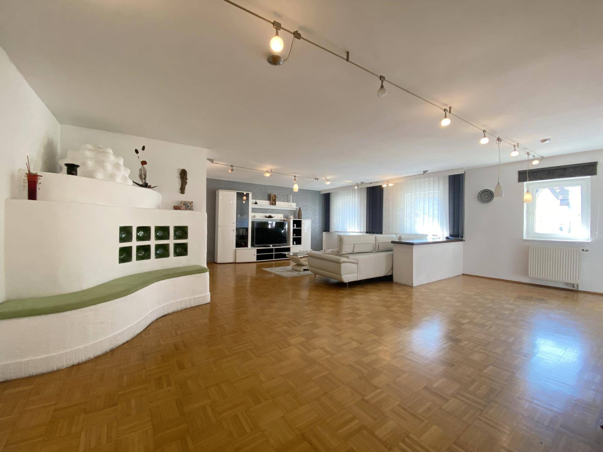 Geräumiges Wohnzimmer mit Parkettboden, weißem Kachelofen und moderner Beleuchtung.