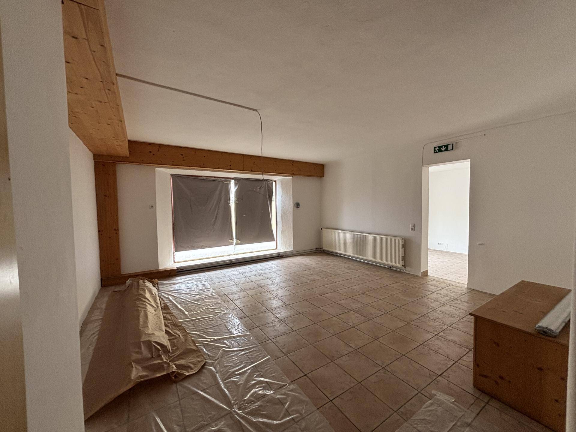 Geräumiger Innenraum mit sichtbaren Holzbalken, Fliesenboden und einem großen Fenster.