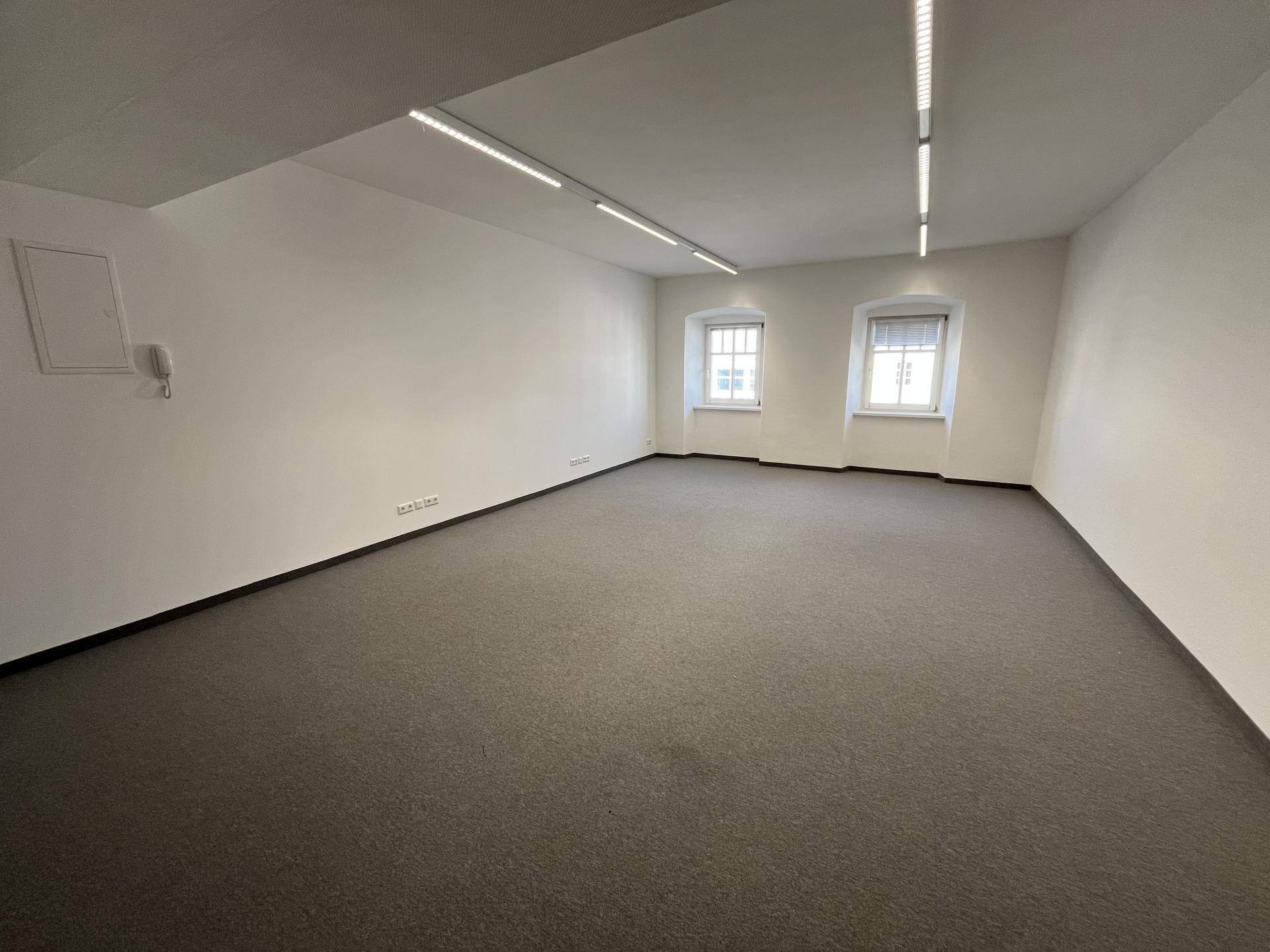 Langer, leerer Büroraum mit dunklem Teppichboden, weißen Wänden und zwei kleinen Fenstern.
