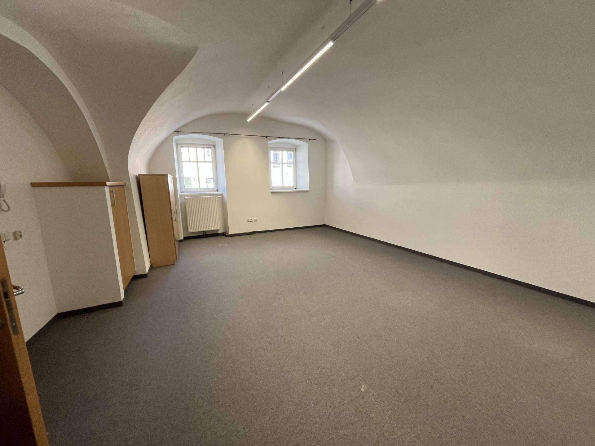 Leerer Raum mit Gewölbedecke, dunklem Teppichboden, kleinen Fenstern und eingebauten Holzschränken.
