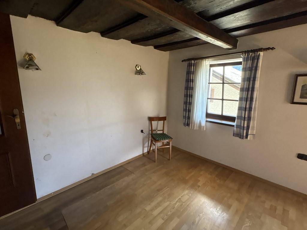 Kleines Zimmer mit Holzboden, sichtbaren Deckenbalken und einem Fenster mit karierten Vorhängen.