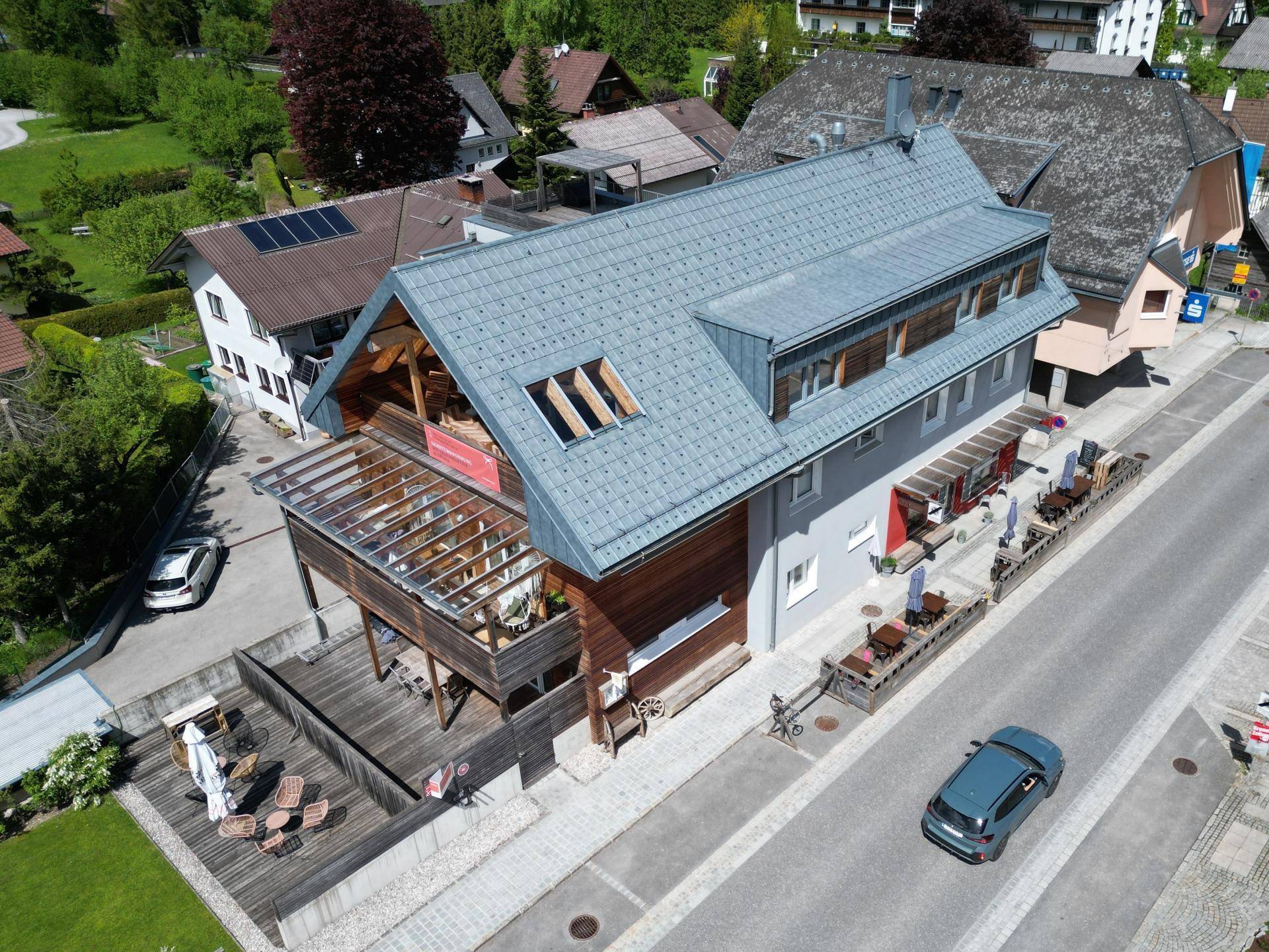Luftaufnahme eines Gebäudes mit Holzfassade, Terrassen und umliegender Bebauung.