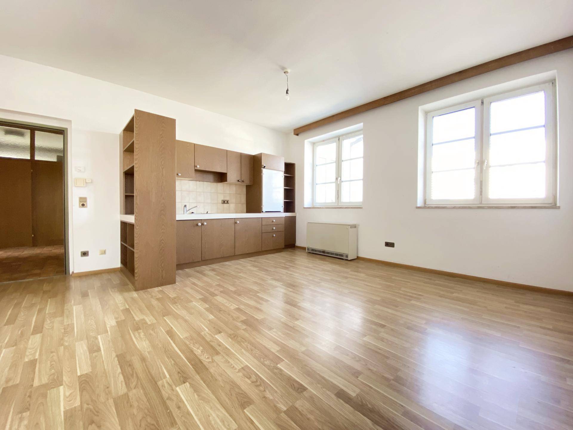 Offener Wohnbereich mit einfacher Küchenzeile, Holzfußboden und Fenstern, die den Raum erhellen.