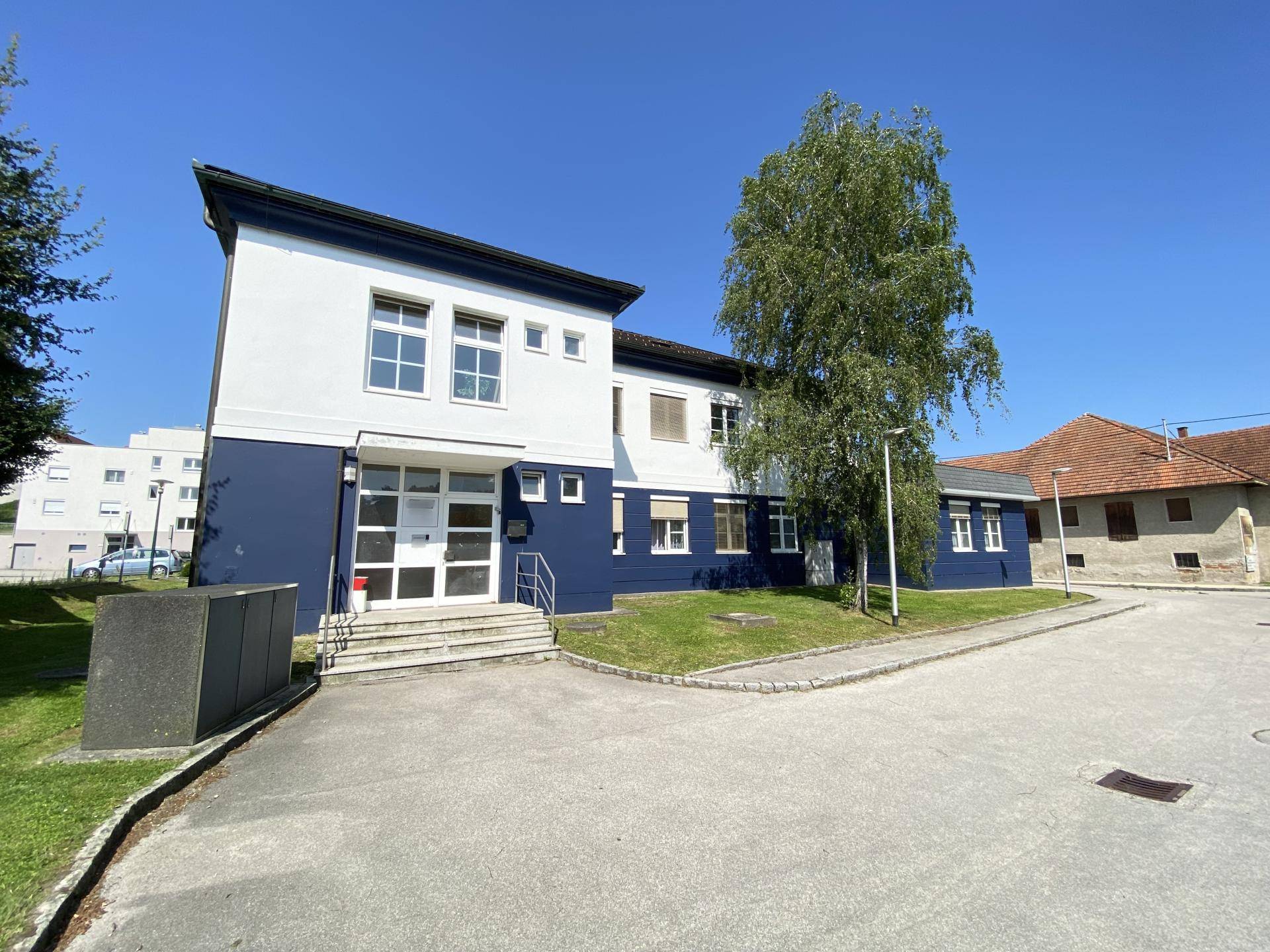 Modernes Gebäude mit blau-weißer Fassade, großen Fenstern und einem gepflegten Eingangsbereich, umgeben von Grün.