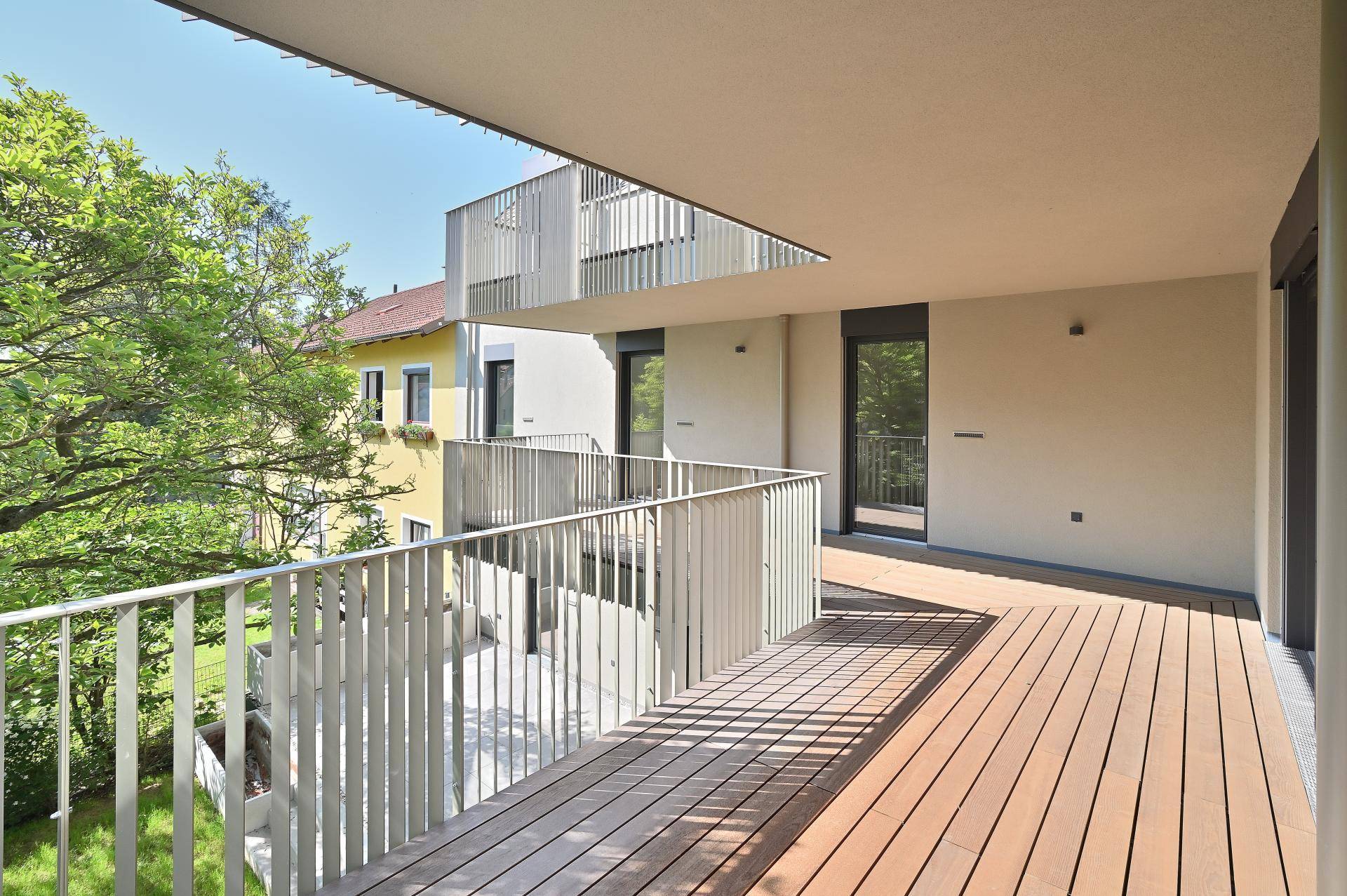 Langer Balkon mit Holzboden und modernem Geländer, umgeben von Bäumen und Häusern.