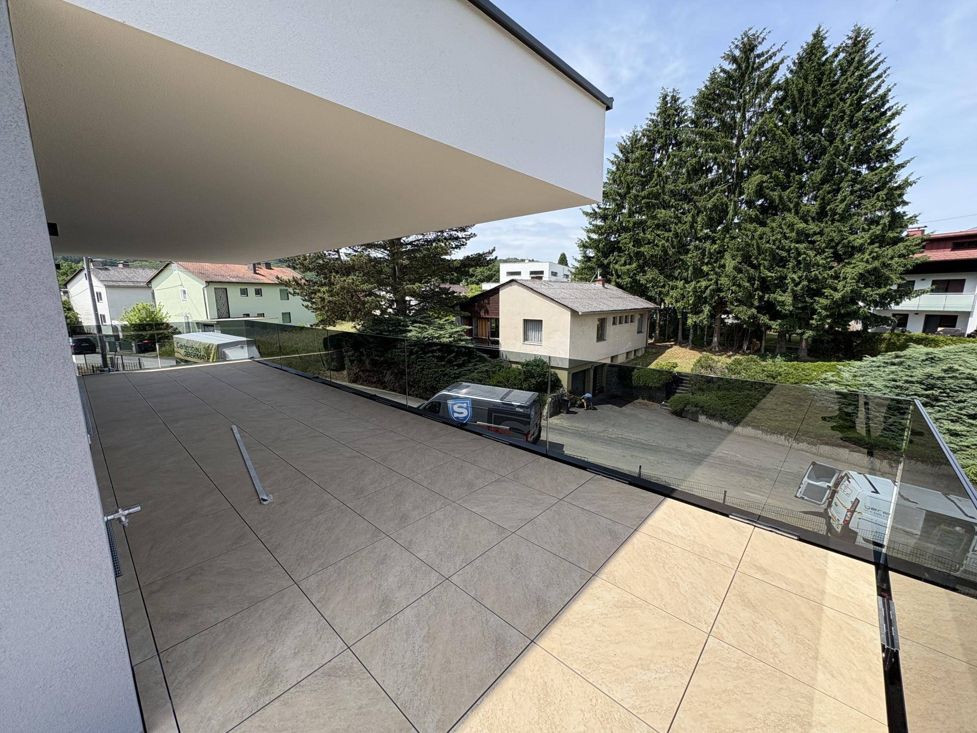 Sonnige Terrasse mit beigen Fliesen und weitem Blick auf grüne Bäume und Nachbarhäuser.