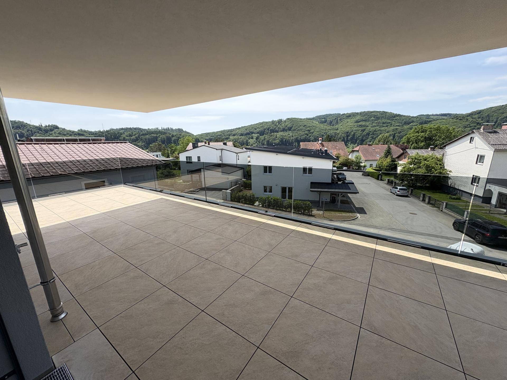 Weitläufige Terrasse mit Glasgeländer und Blick auf die umliegende Wohnsiedlung und grüne Hügel.