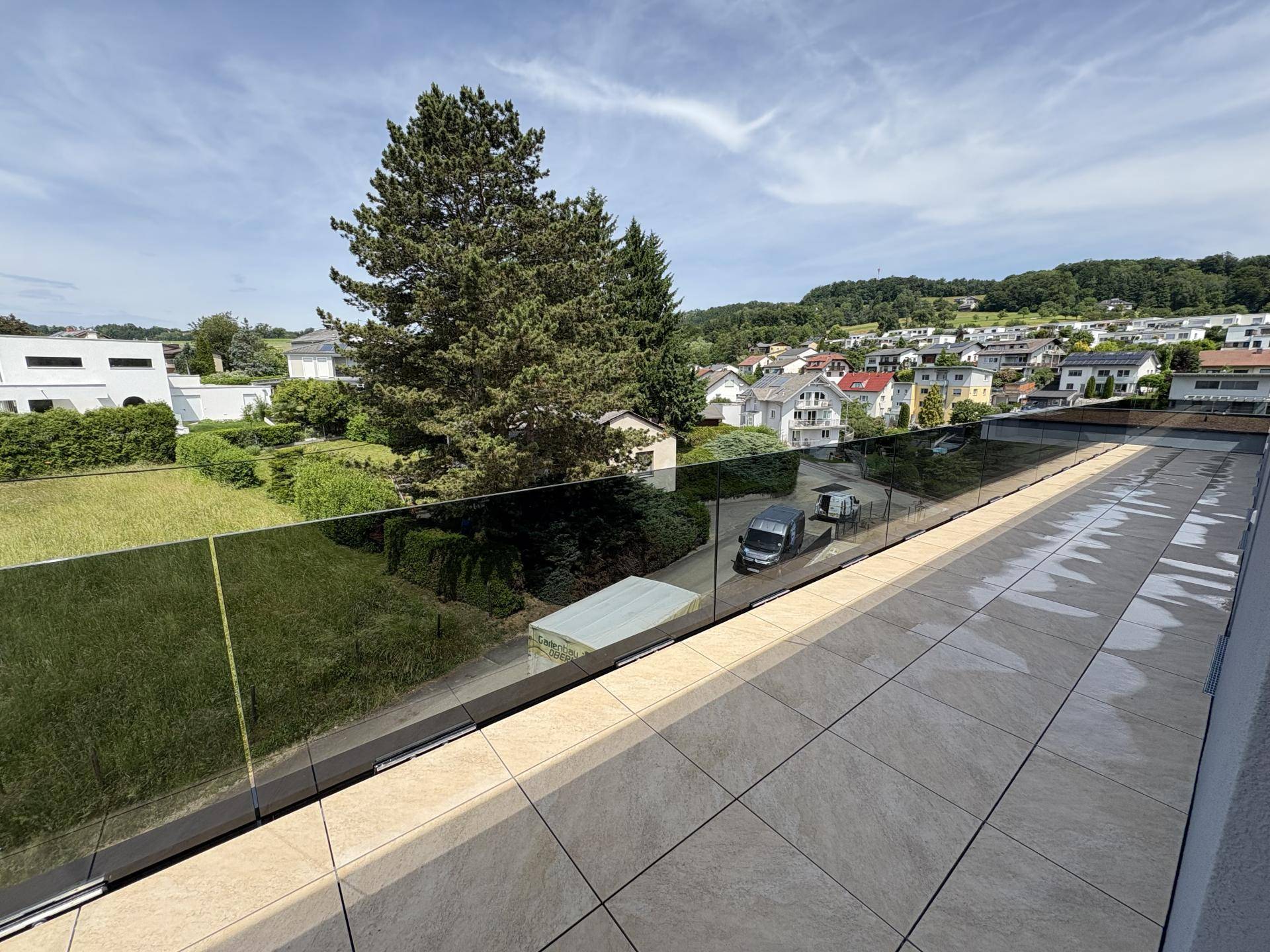 Weitläufige Dachterrasse mit modernen Fliesen und Glasgeländer, umgeben von Grünflächen und Bäumen.