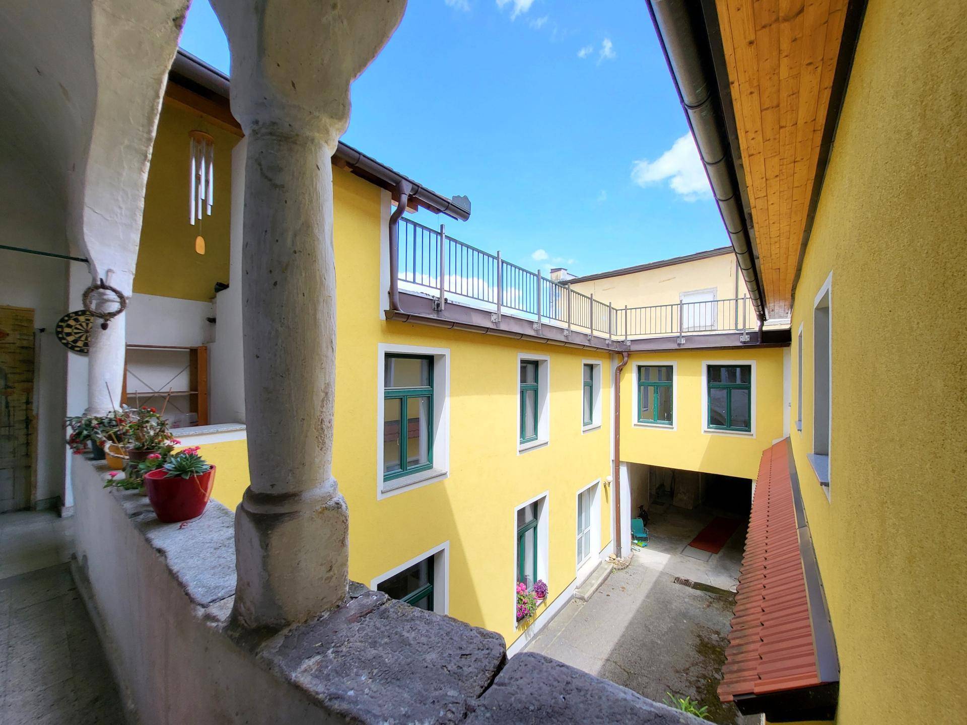 Blick in einen sonnigen Innenhof mit gelben Fassaden und grünen Fensterrahmen von einem überdachten Gang.