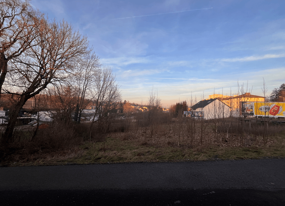 Blick auf ein unbebautes Grundstück bei Sonnenuntergang mit kahlen Bäumen und Gebäuden im Hintergrund.