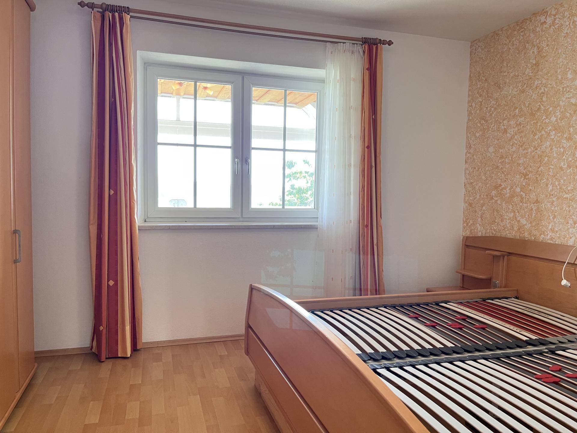 Schlafzimmer mit Holzbettrahmen, Laminatboden und Fenster mit Blick ins Grüne.
