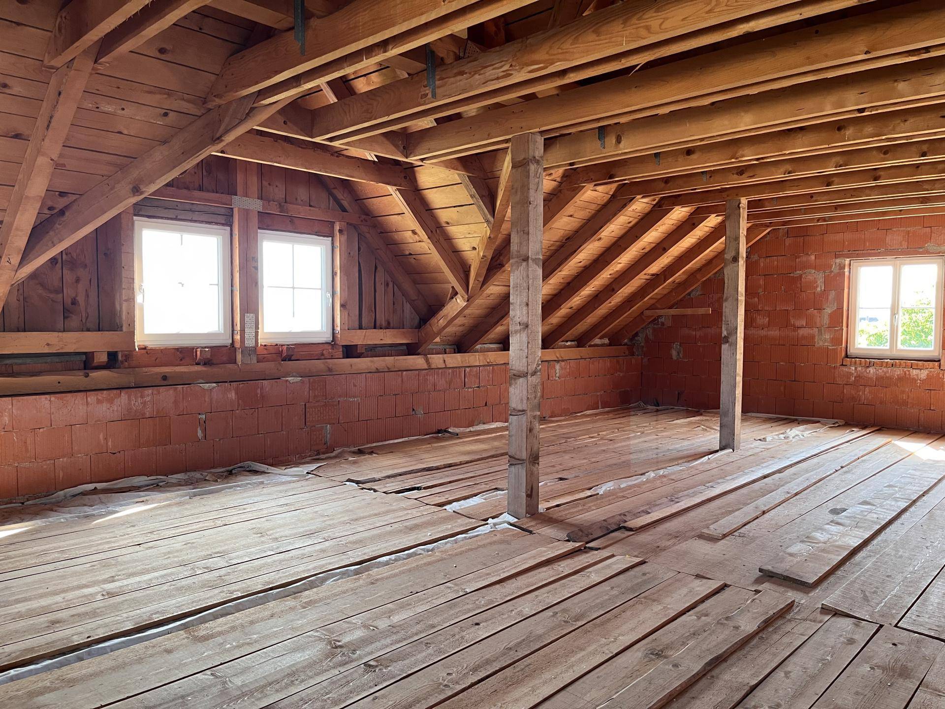 Dachboden im Rohbau mit Holzbalken, Holzboden und Fenstern.