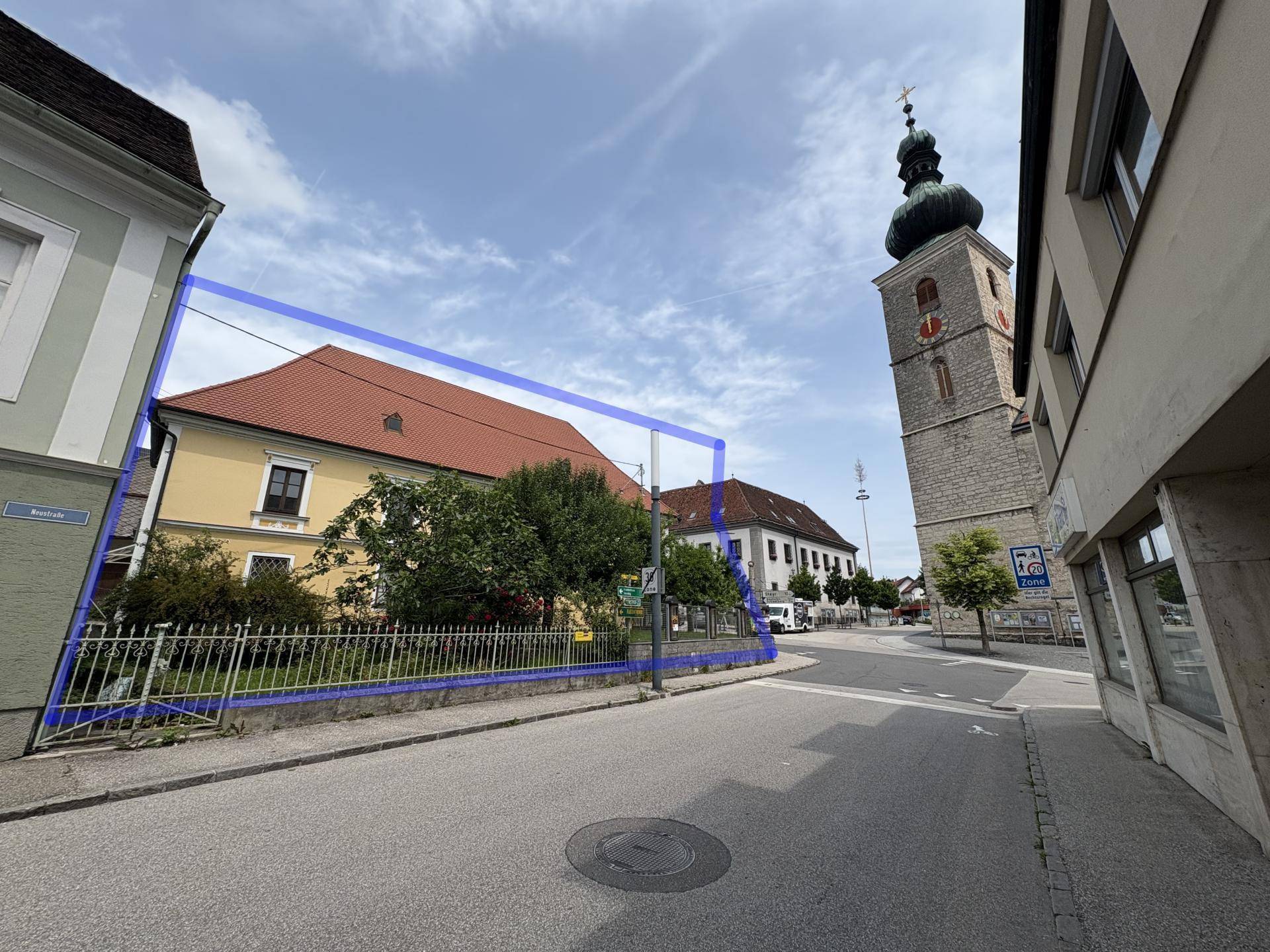 Gelbes historisches Gebäude mit blauem Grundstücksumriss und einem Kirchturm im Hintergrund.