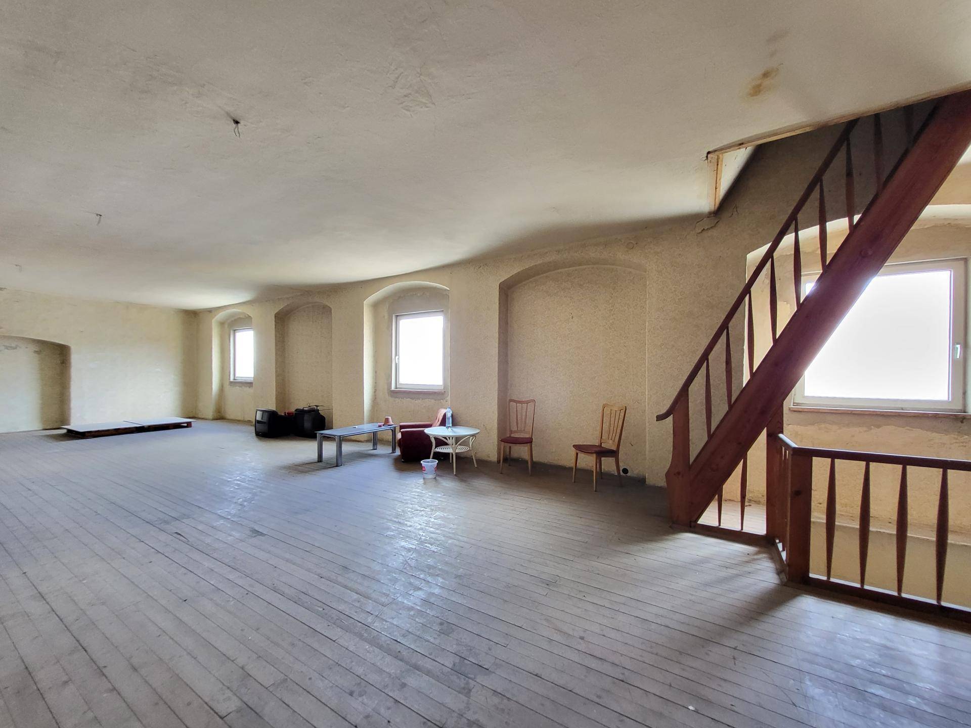Großer, leerer Raum mit Holzdielenboden, gewölbten Fenstern und einer Holztreppe.