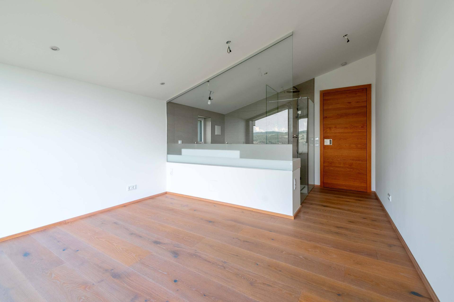 Helles Schlafzimmer mit Holzboden und integriertem Badezimmerbereich, abgetrennt durch eine Glaswand.