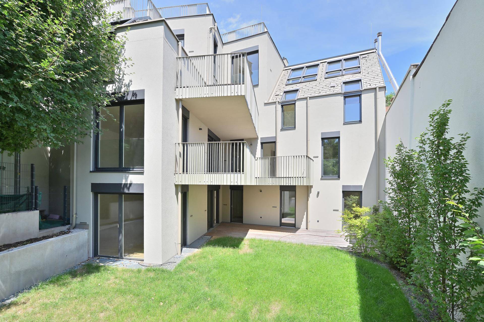 Rückansicht des modernen Gebäudes mit mehreren Balkonen, Dachfenstern und einem kleinen Garten mit Terrasse.