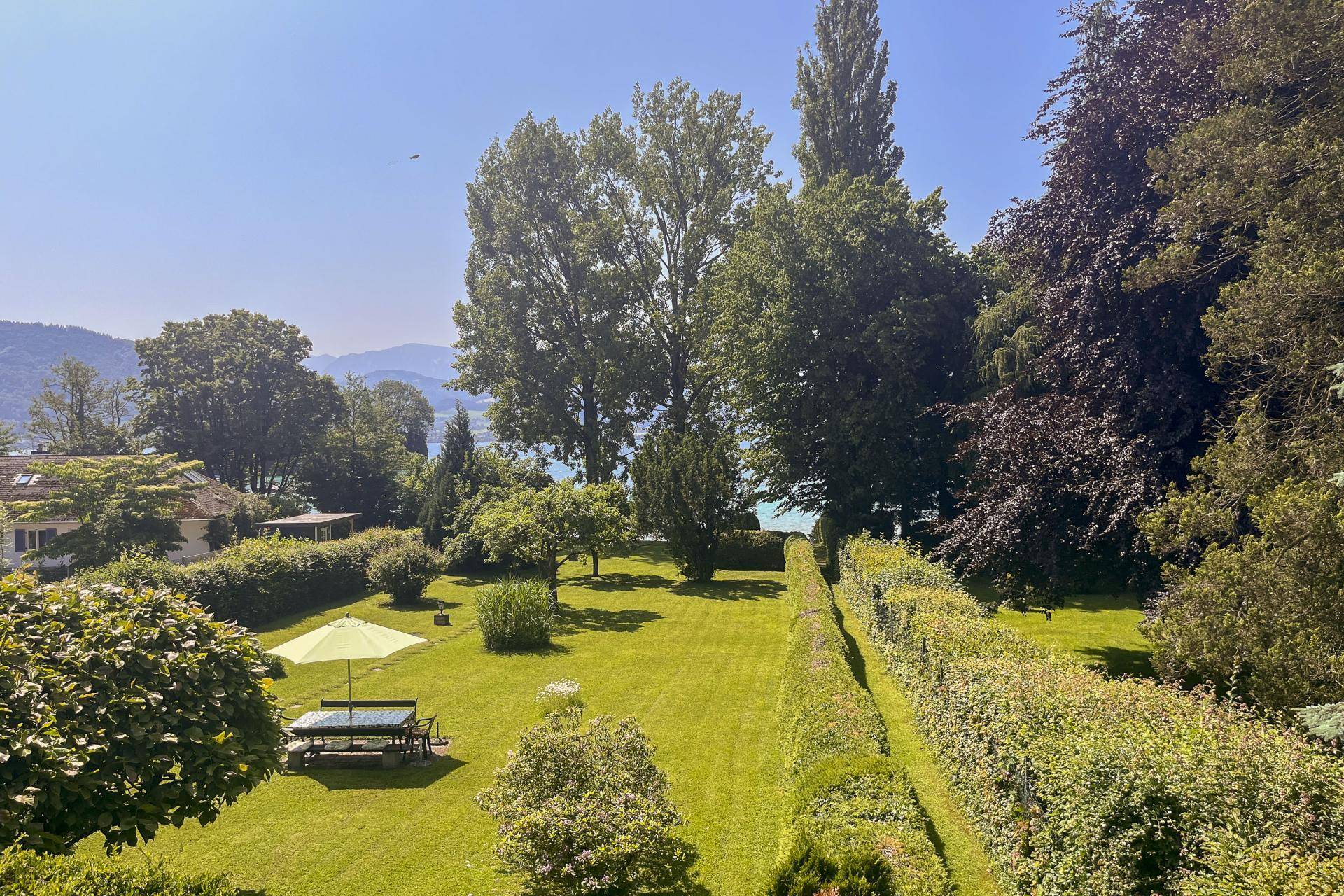 Weitläufiger, gepflegter Garten mit Rasen, hohen Bäumen und Blick auf den See.