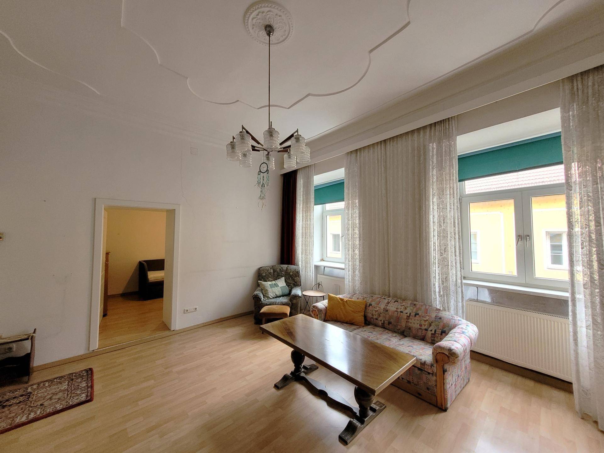 Geräumiges Wohnzimmer mit Holzboden, klassischem Sofa und großen Fenstern, die viel Licht hereinlassen.