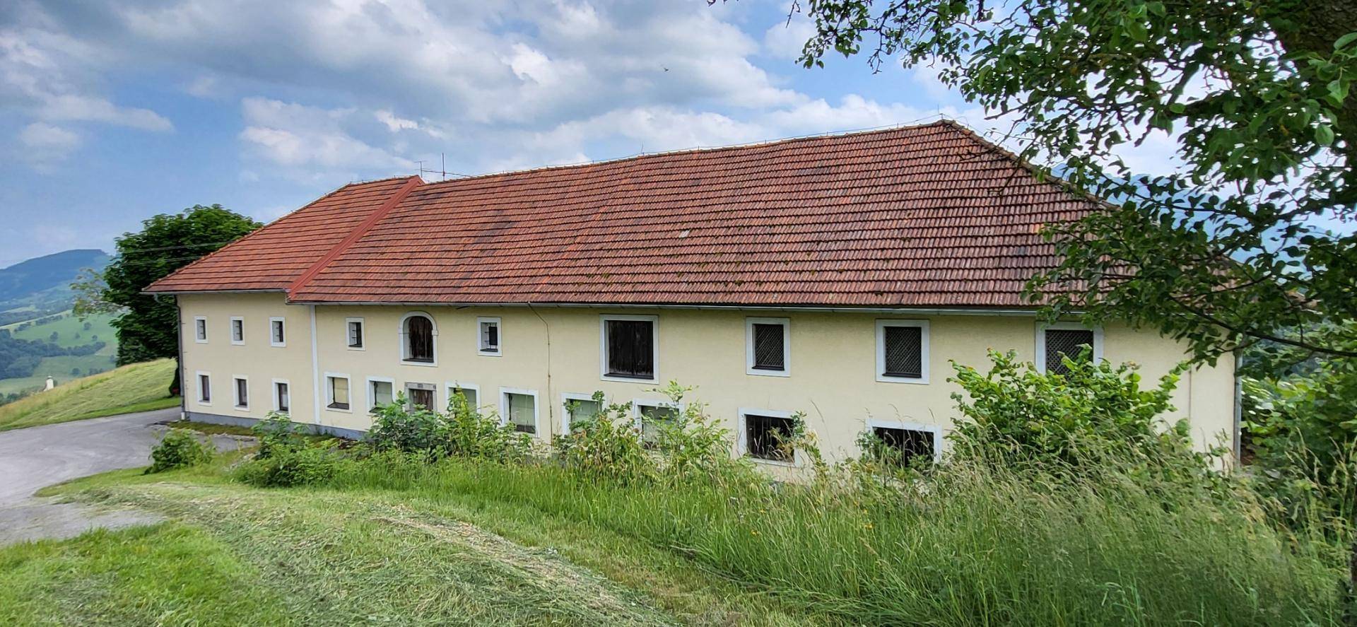 Langes, traditionelles Gebäude mit hellgelber Fassade und rotem Ziegeldach, umgeben von viel Grün.