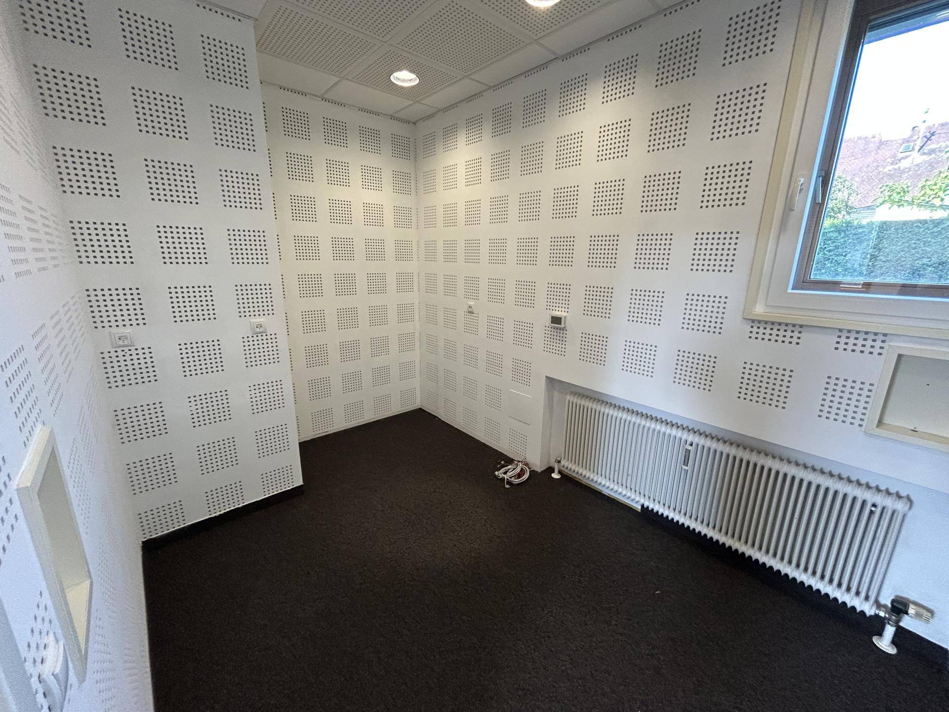 Kleines Büro mit schallabsorbierenden Akustikpaneelen an den Wänden, Teppichboden und Fenster.